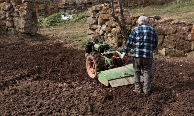Elderly man using a green tiller in a rustic field with stone walls, embodying traditional farming. - Photo by Zakhar Vozhdaienko on Pexels